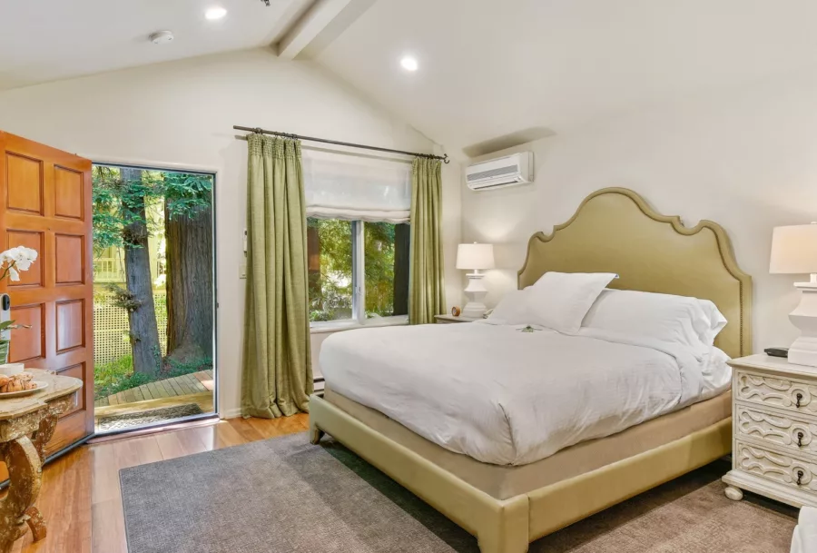 Plush king bed, bedside tables and lamps in a room with a peaked ceiling, with door open to trees