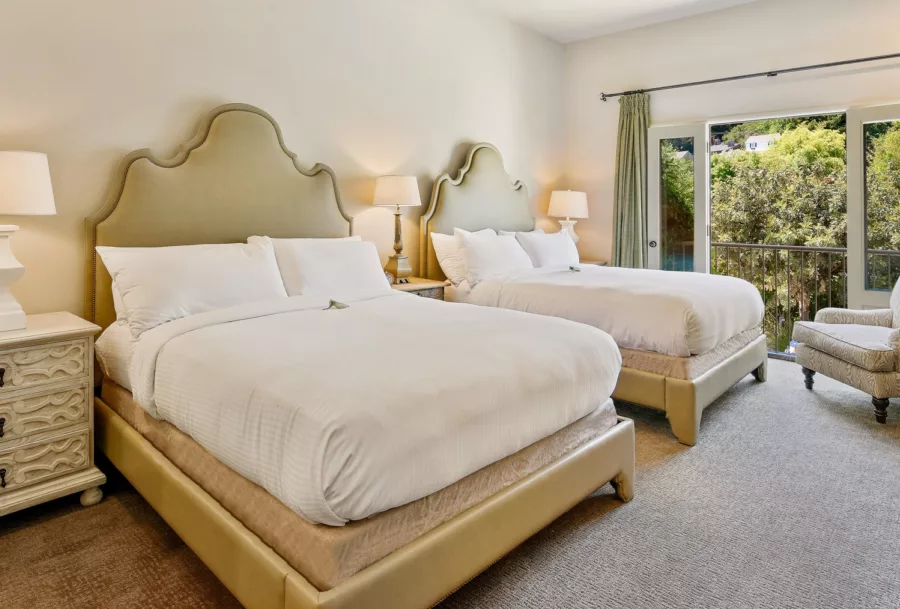 Two plush queen beds with fabric headboards, side tables and lamps, side chair, and large glass doors open to trees